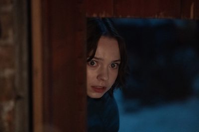 A woman looks in fear hiding behind a wall.