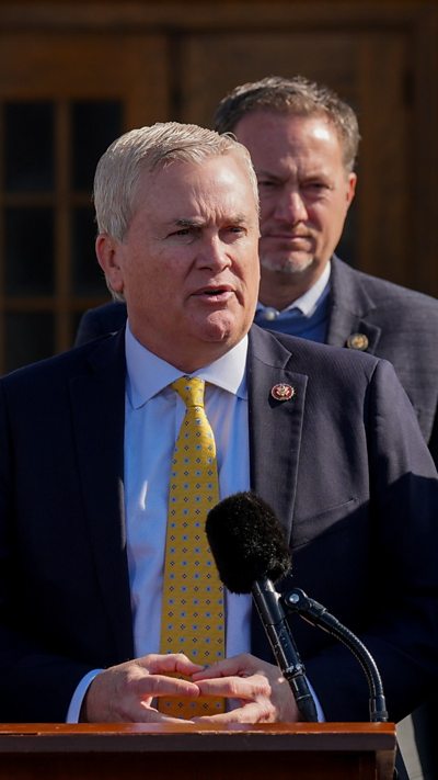 A man wearing a yellow tie and blue suit speaks at a microphone.