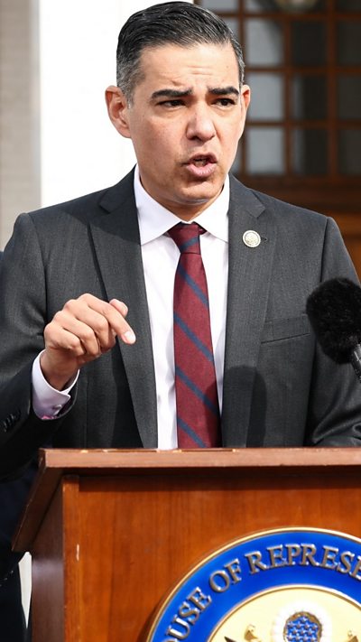 A man wearing a dark suit and tie speaks into a microphone.