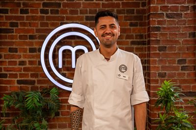 A man in the MasterChef studios with chef whites poses - he has a SEB name badge. 