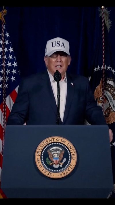 Donald Trump standing in front of a podium, wearing a white hat which says USA on it
