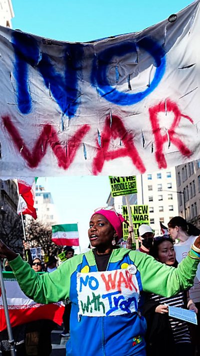 A woman wearing a colourful sweater holds up a banner that says "No War" as other protesters march behind her.