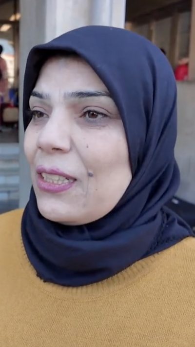 A woman wearing a yellow top and a blue headscarf, speaking to the camera