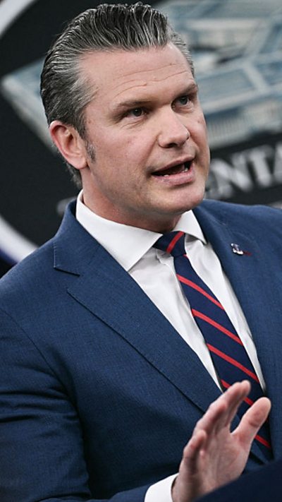 A man wears a blue suit and red striped tie while speaking at a microphone at the Pentagon.