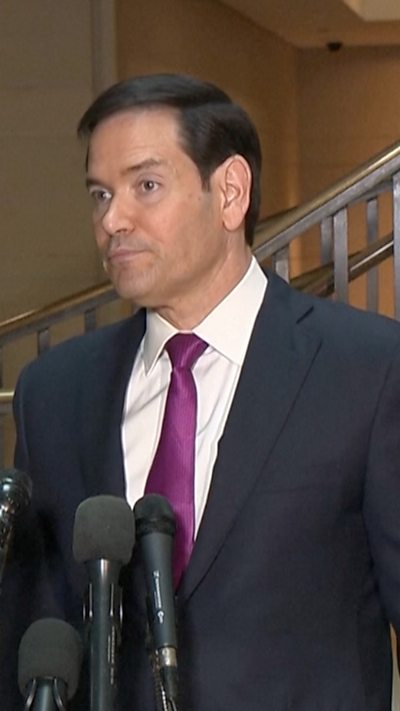 Marco Rubio standing in front of microphones wearing a black suit and white shirt with a purple tie.