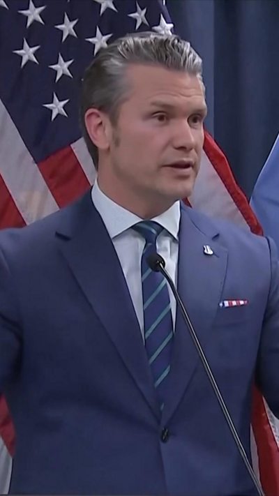 Pete Hegseth wearing navy suit and standing in front of US flag