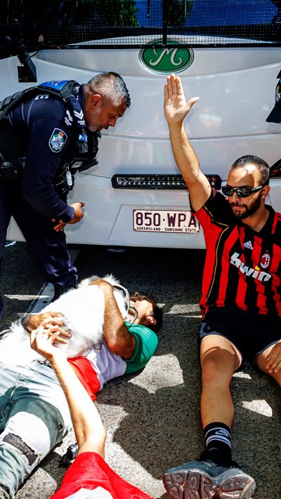 A man lies in front of a bus as another man sits next to him and a police man stands looking down at them.