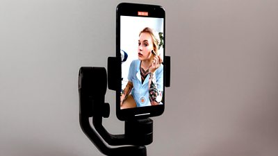 A shot of a mobile phone in a stand, the screen is facing towards us, and on the phone screen is someone giving a makeup tutorial.