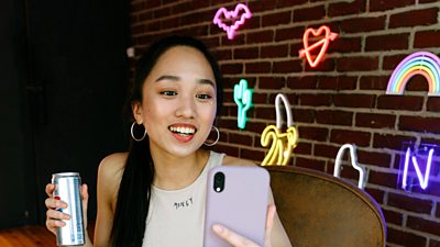 An influencer presents into a mobile phone they are holding in their hand. They are seated in a room or studio lit with bright neon signs and lights.