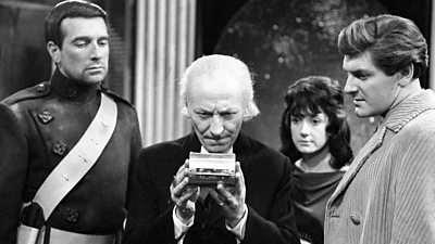 Black and white still from Doctor Who episode The Devil's Planet, featuring William Hartnell as the Doctor, surrounded by three people as he looks at something in his hands intently.