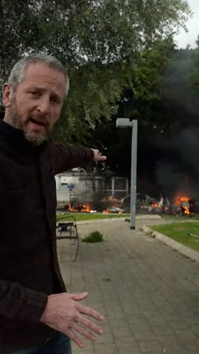 Jon Donnison looks at the camera while pointing towards a fire after a strike in Tel Aviv