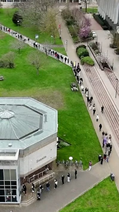 A drone shot shows a long single-file queue winding round a path towards a university building