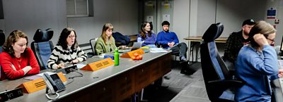 Group seated around a studio table with scripts, laptops and microphones during a recording session.