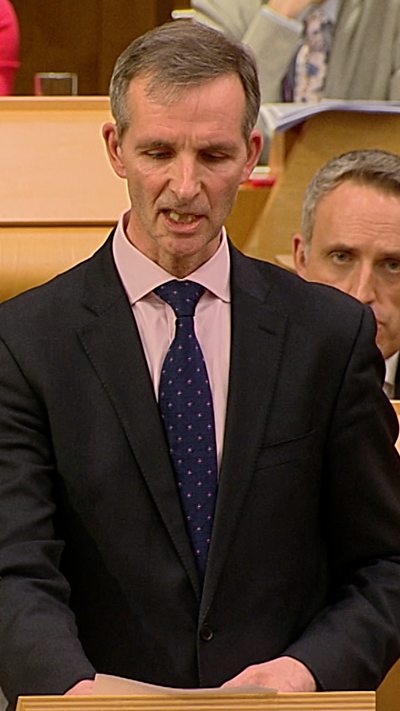Man in a dark suit and tie in the Scottish Parliament chamber