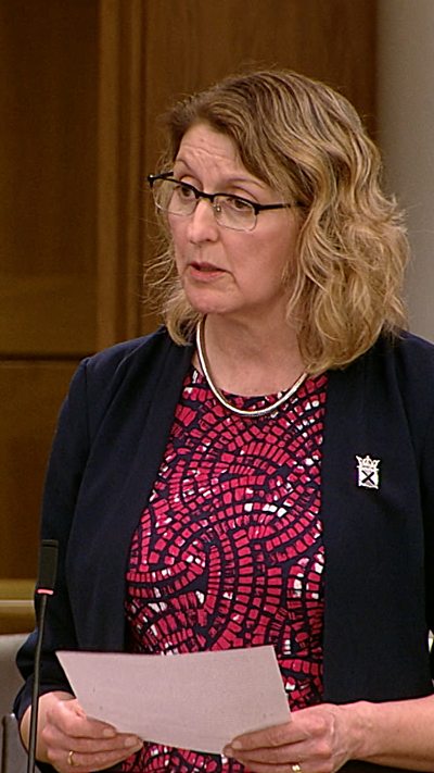 Woman in dark jacket and patterned top in the Scottish Parliament chamber.