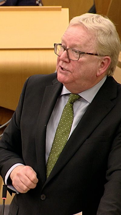 A man in a dark suit and green tie in the Scottish Parliament chamber.