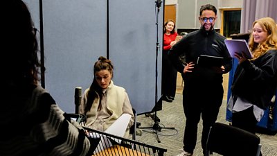 People in a studio setting, one seated at a microphone while others stand nearby holding scripts, with screens and lighting equipment visible.