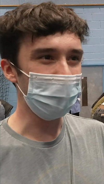 A young man with brown hair and a face mask on speaks to the camera