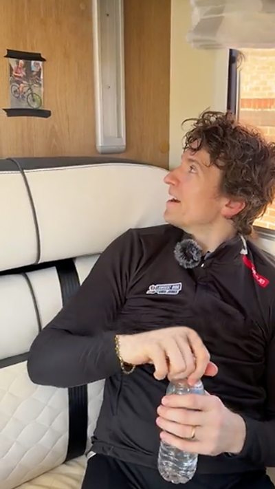 Greg James, wearing a black cycling top, is sat looking to the side of a picture of his dad which is stuck to the wall