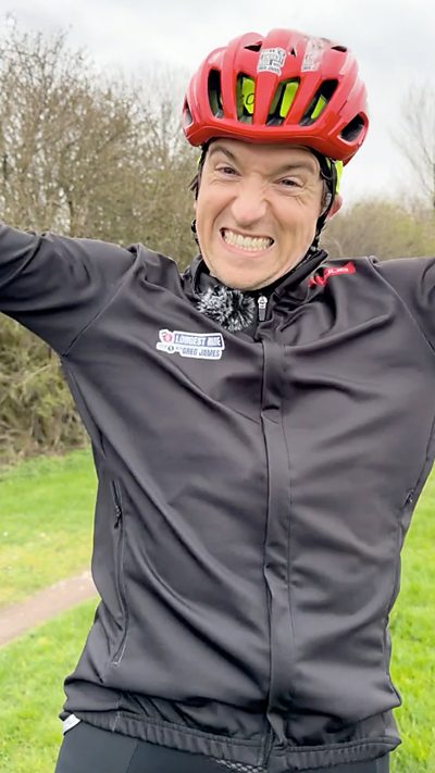 Greg James, wearing a black cycling top and red helmet, celebrates