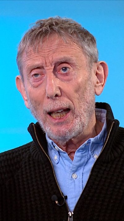 Michael Rosen in a blue shirt and zip up jumper in front of a blue screen