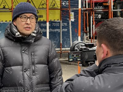 A man speaks to a camera. He's wearing a black puffy jacket and standing in front of a construction site.