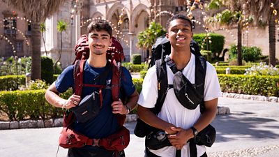Two young men stand in the grounds of a cathedral in sunny Sicily wearing large backpacks. They smile to camera.