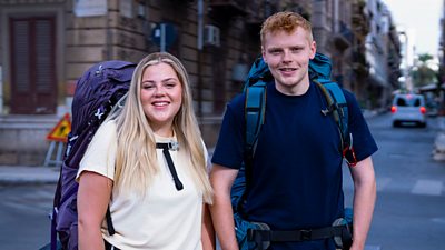 A young woman and young man stand together on a street in Palermo, Sicily, wearing backpacks and smiling to camera.