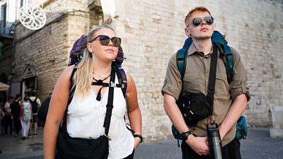 A young woman and man stand together against the backdrop of a large stone wall. They both wear sunglasses and backpacks as they look up at something off camera