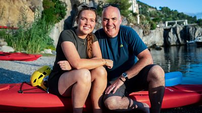 A young woman and a man sit together on a red kayak. They smile to camera with sunglasses on their heads.