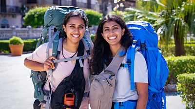 Two young women smile to camera in the sunny grounds of a cathedral. They both carry large backpacks.