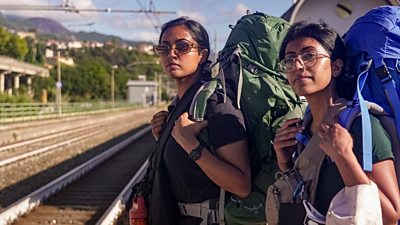 Two young women stand poised to move on a train platform, both holding their backpacks as they look at something off screen