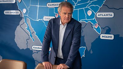 A man in a suit stands against a map of north and south America, leaning on a chair