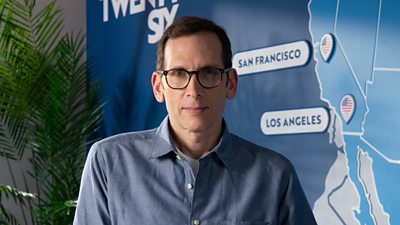 A man in a blue shirt and glasses looks to camera, standing against the backdrop of a map of US cities