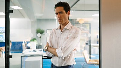 A man in a white striped shirt stands in an office with his arms folded, looking warily to camera