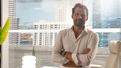A man in a white shirt sits with his arms folded, smiling to camera, against the backdrop of a sunny skyline