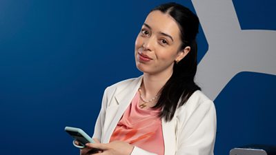 A woman in a white blazer and peach top smiles to camera, holding a mobile phone