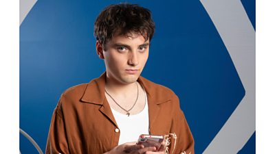 A man in a brown shirt and white top looks to camera, holding a mobile phone