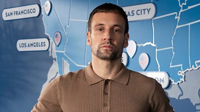 A man in a brown polo shirt looks to camera, standing against the backdrop of a map of north American cities