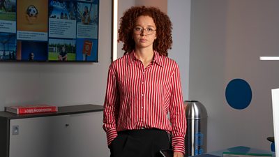 A woman with curly red hair and glasses stands in an office, wearing a white and red striped shirt