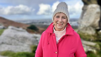 A woman in a pink coat, cream jumper and cream hat smiles to camera, standing on a rocky landscape