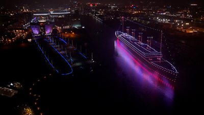 An aerial night view of a brightly illuminated ship outlined in colourful lights, floating on dark water with its pink and purple reflection beneath it. To the left, a waterfront area and buildings are also lit with blue and purple lighting, while the city skyline in lit up in the distance