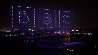 Three large illuminated drone formations spell out the BBC in blocks in the night sky above a waterfront cityscape