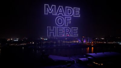 Nighttime aerial view of a waterfront with a large drone light display forming the words “MADE OF HERE” in bright white lights above the city. Buildings and their reflections are visible below