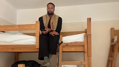 A man sits at the top of a bunk bed, smiling with his hands crossed