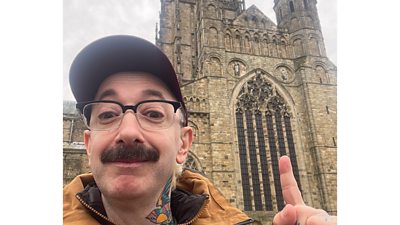 A man in a baseball hat and glasses smiles as he takes a selfie outside a cathedral, pointing up to its tall spire