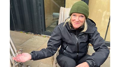 A woman in a wooly hat and dark jacket smiles as she sits in a chair with a small bird perched on her outstretched hand