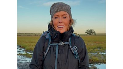 A woman in a wooly hat and warm jacket smiles as she stands in a waterlogged field