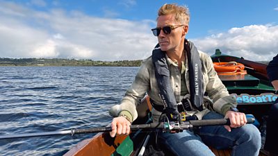 A man sits in a boat holding a fishing pole. He wears a shirt, jeans and a life jacket.
