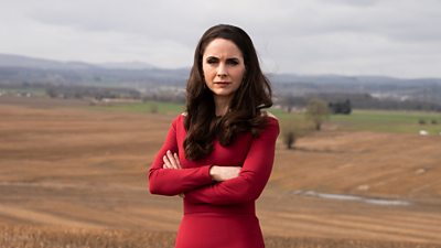 Photo of Laura Fraser in character as Cat. Wearing a formal red dress, she stands in a field. The countryside stretches into the horizon behind her. 